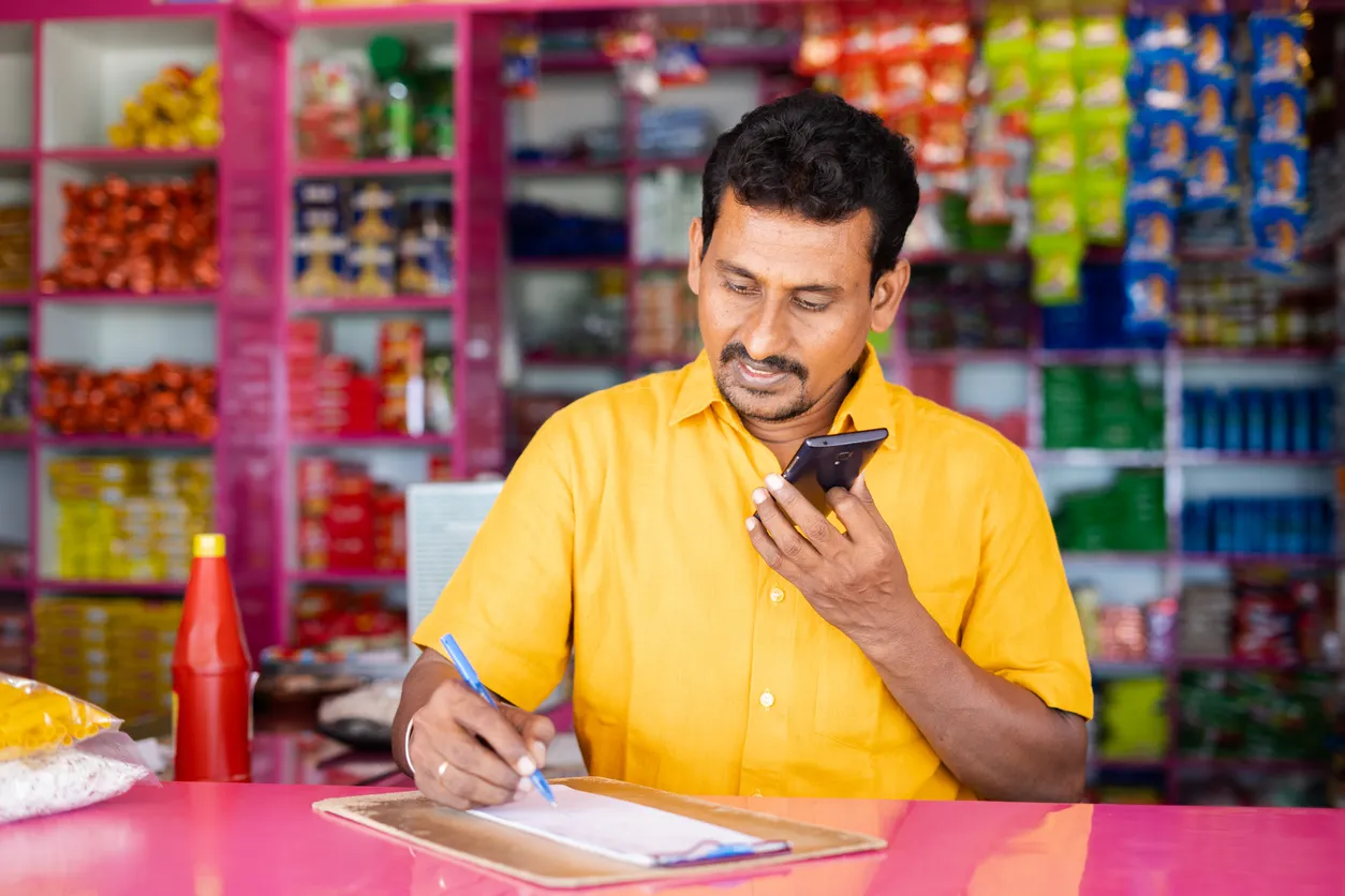 Un hombre de negocios de comestibles anota los pedidos mientras habla con un cliente por teléfono móvil de media gama