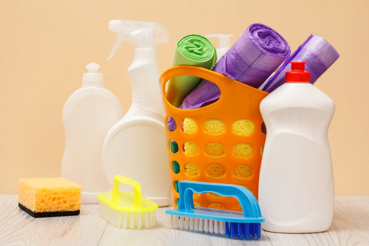 Botellas de plástico de limpiador de vidrios y azulejos, cesta con bolsas de basura, esponjas y cepillo sobre fondo beige. 
