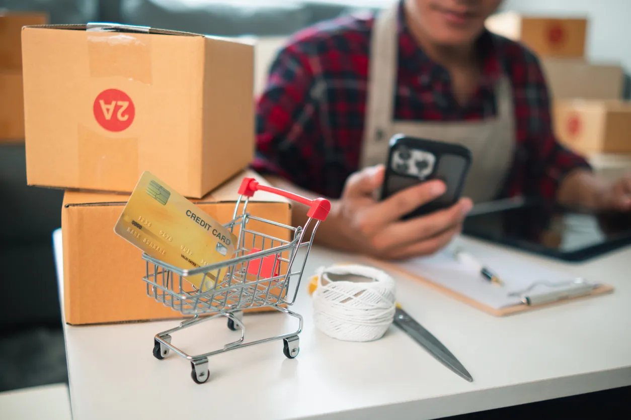 Hombre sentado con overoll revisando su celulkar y  con cajas y un carrito de supermercado represantanod las ventas online