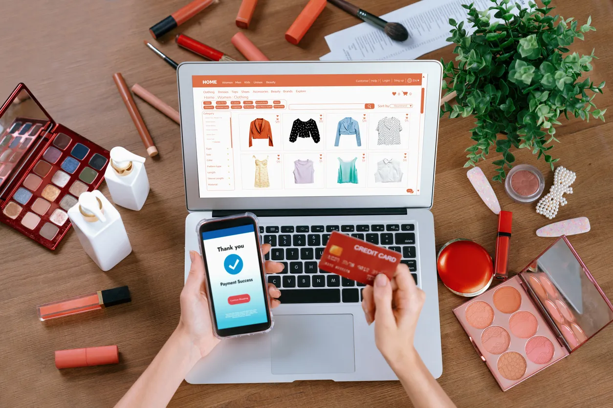 Manos de mjuer con un celular y una tarjeta bancaria frente a una laptop con una tienda de ropa online en la pantalla