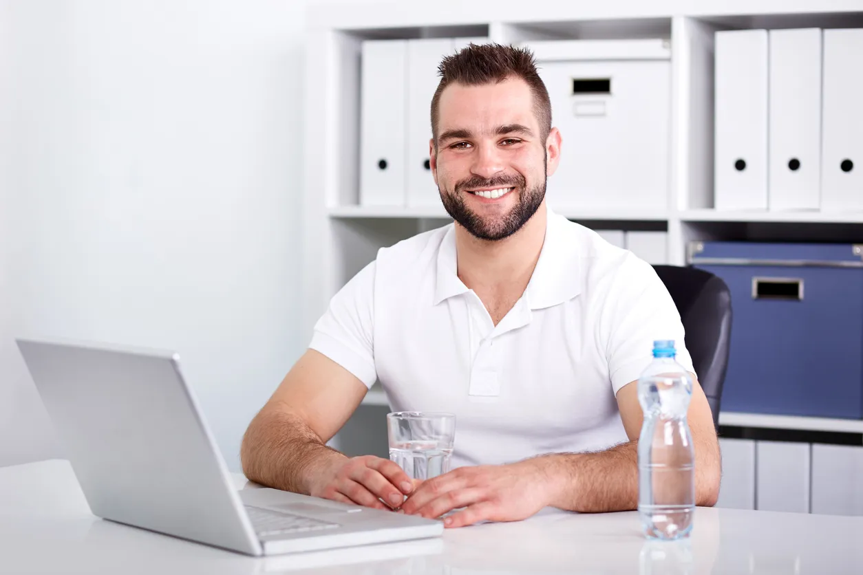 Joven con playera blanca con cuello sentado y con un vaso de agua y laptop sobre su escritorio. 