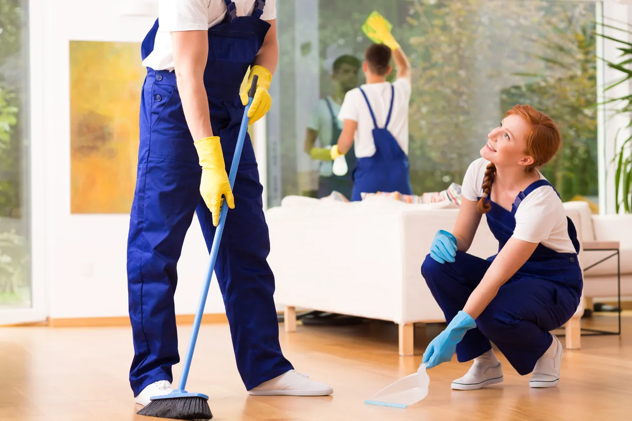 Jovenes relizando la limpieza de la sala de una casa moderna con overoles, playera blanca y guantes. 