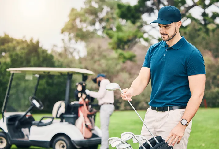 Hombre con playera y gorra azul sosteniendo un palo de golf, y al fondo un carrito de golf