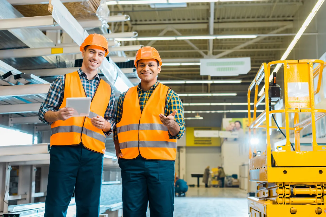 Dos trabajadores jóvenes con chaleco y cascos de seguridad naranja en una gran nave industria, usan coolers industriales