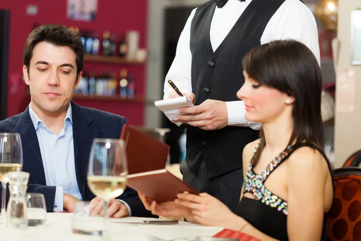 Pareja sentada en un restauratnte y siendo atendidos por un mesero con chaleco negro