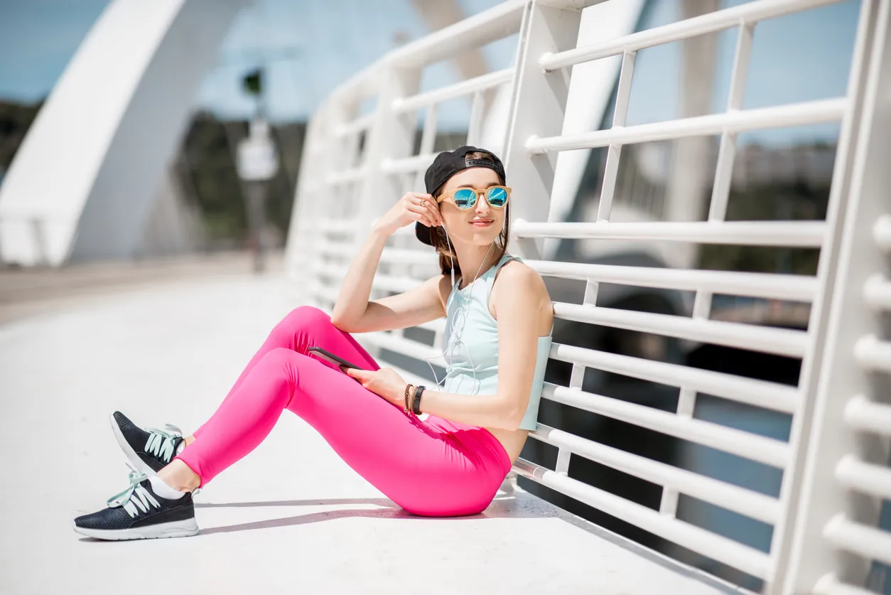 mujer joven con ropa deportiva, lentes de sol, audífonos sentada en el suelo recargada sobre un puente