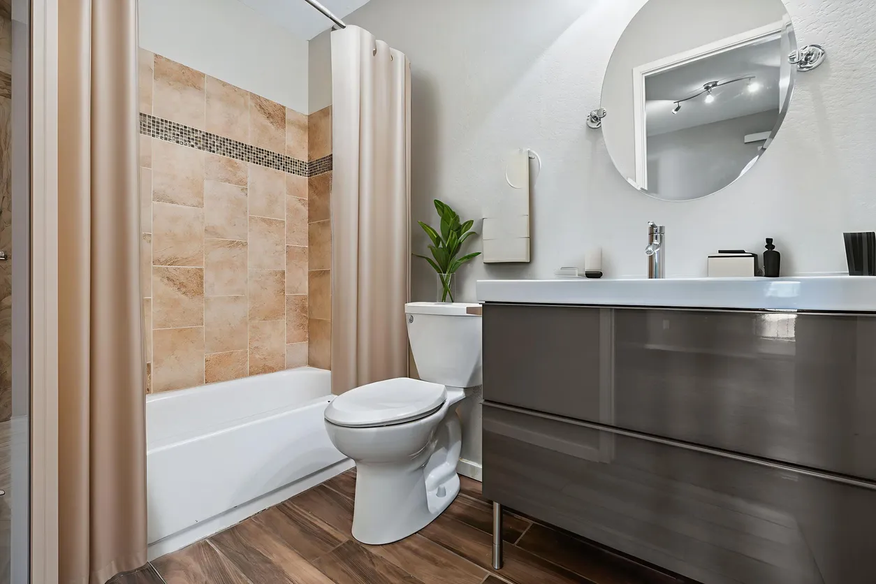 Baño moderno con gabiente para baño con lavabo y gran espejo redondo..Taza y un tina al fondo. 