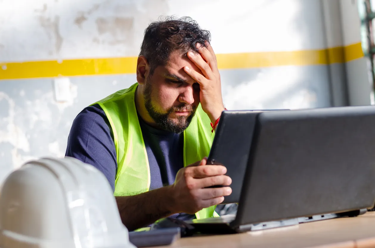 trabajador con chaleco de seguridad sentado en su escritorio mostrando preocupación con una mano en la cabeza