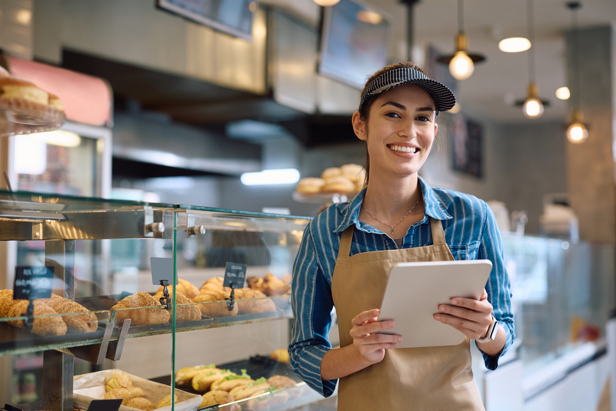 Joven mujer feliz trabajando en una tableta digital en una panadería y mirando a la cámara
