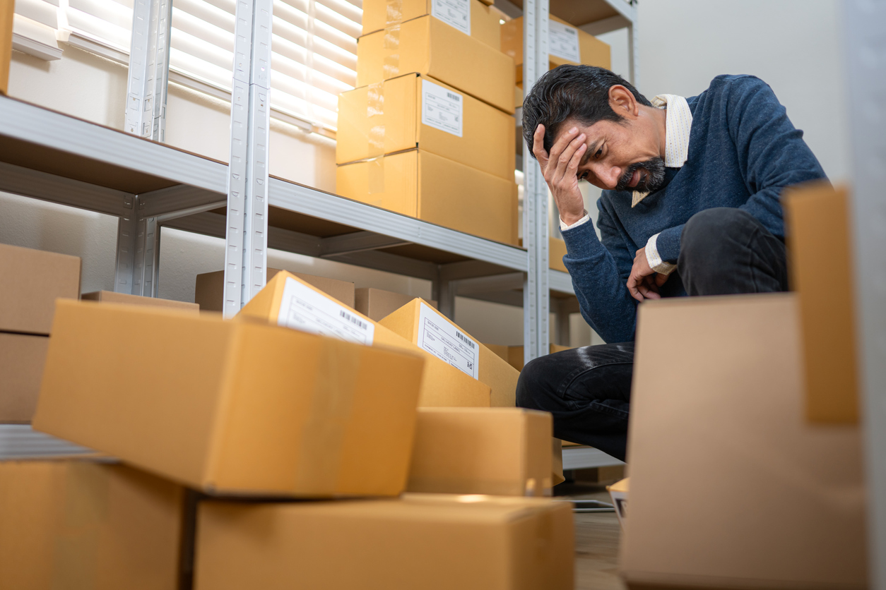 Trabajador de almacén estresado sentado entre cajas de cartón, expresando un problema empresarial 