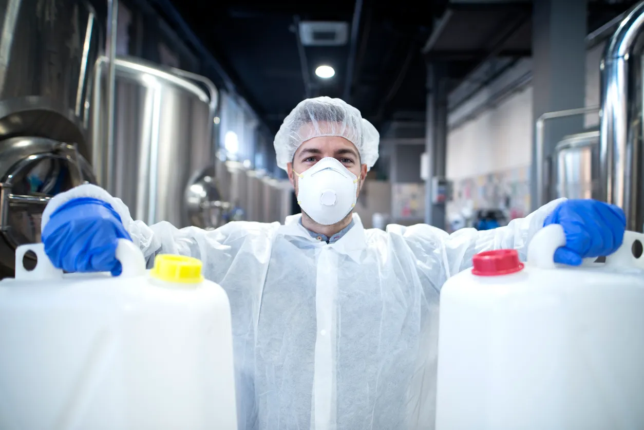 Trabajador industrial con máscara protectora y uniforme blanco sosteniendo bidones de plástico en industria de desengrasantes