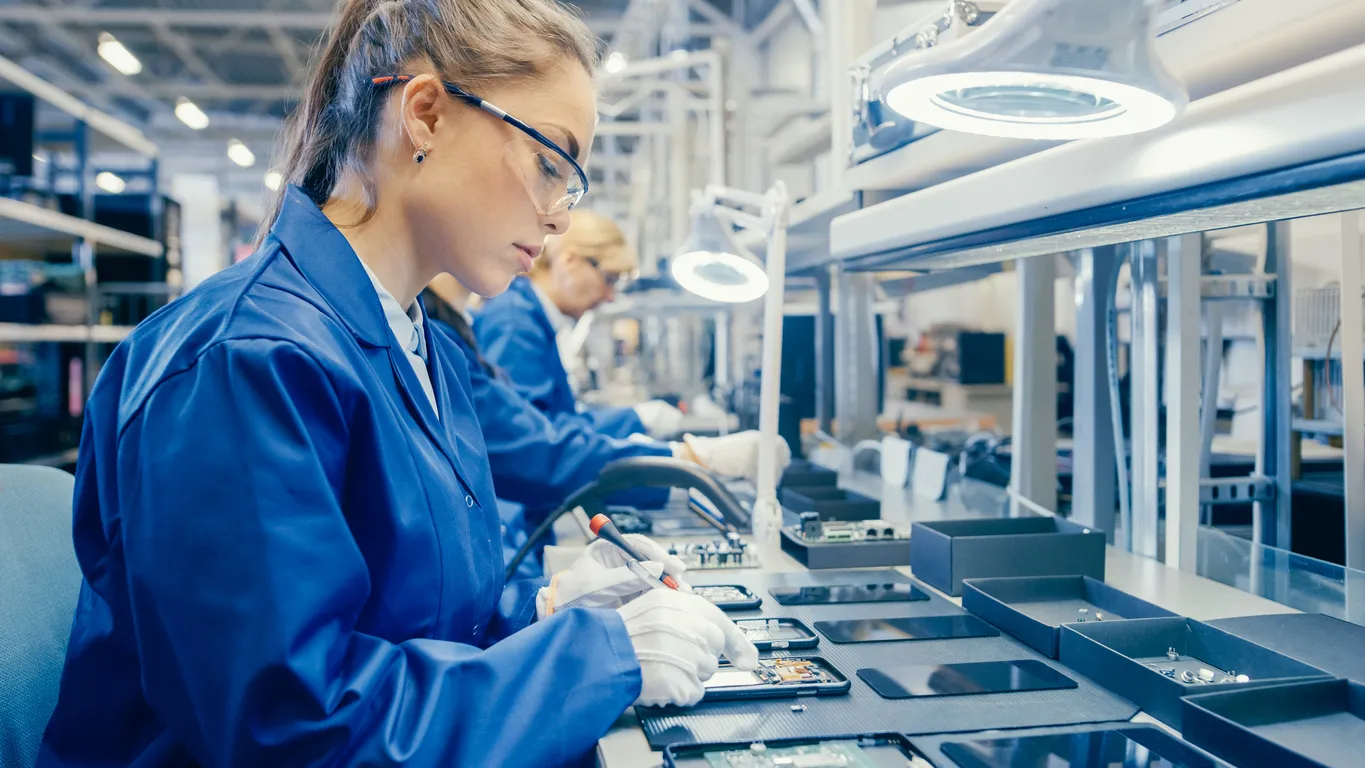 Mujer Electronics fábrica trabajador en azul capa de trabajo y gafas protectoras es ensamblar smartphones con destornillado