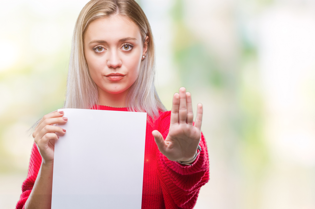 Young blonde woman holding blank paper sheet over isolated background with open hand doing stop sign with serious and confide