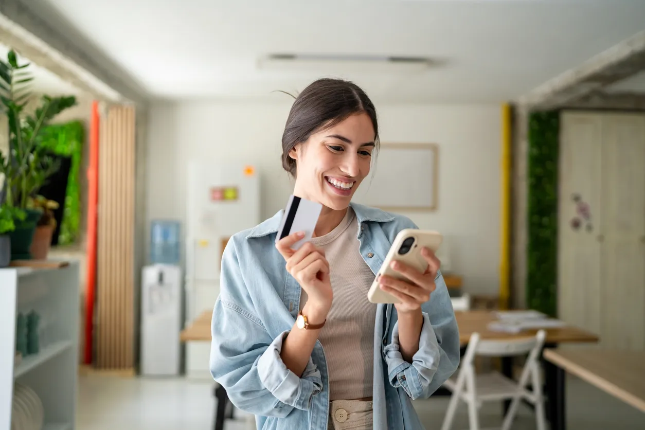 Mujer joven con tarjeta de crédito y teléfono inteligente sonriendo en casa 