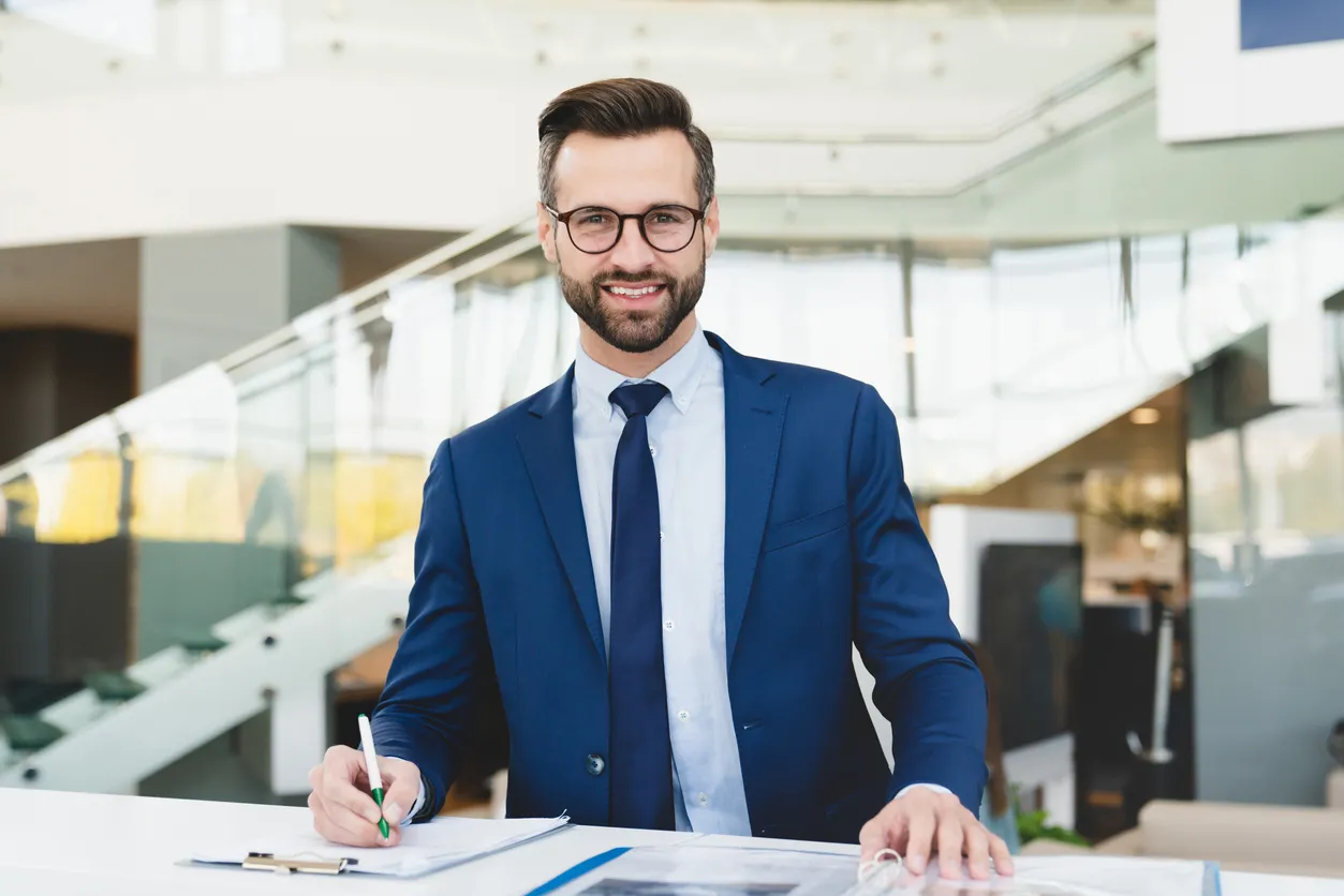 Exitoso hombre caucásico sonriente asistente de tienda recepcionista con atuendo formal escribiendo mirando a la cámara mient