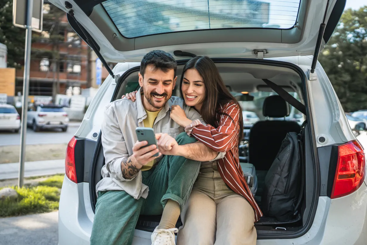 Pareja de adultos jóvenes se sienta en el maletero del automóvil y usa el teléfono móvil en la calle 