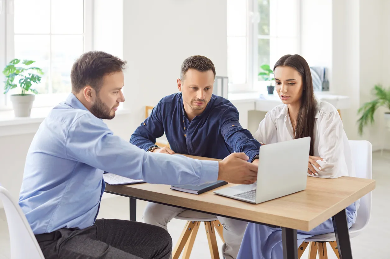 Pareja joven sentada con un agente inmobiliario, explorando opciones inmobiliarias en la pantalla del portátil para comprar 