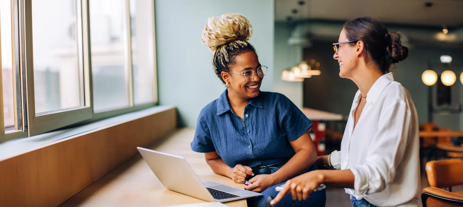 Dos mujeres jóvenes sonriendo y hablando mientras trabajan juntas en una computadora portátil