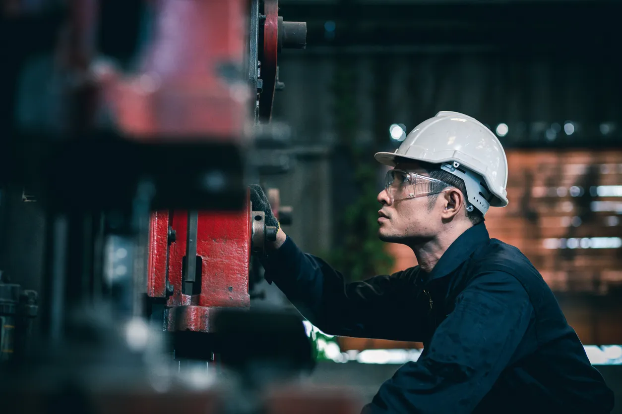 Ingeniero industrial de hombres que lleva un casco blanco mientras está de pie en una fábrica industrial pesada detrás.