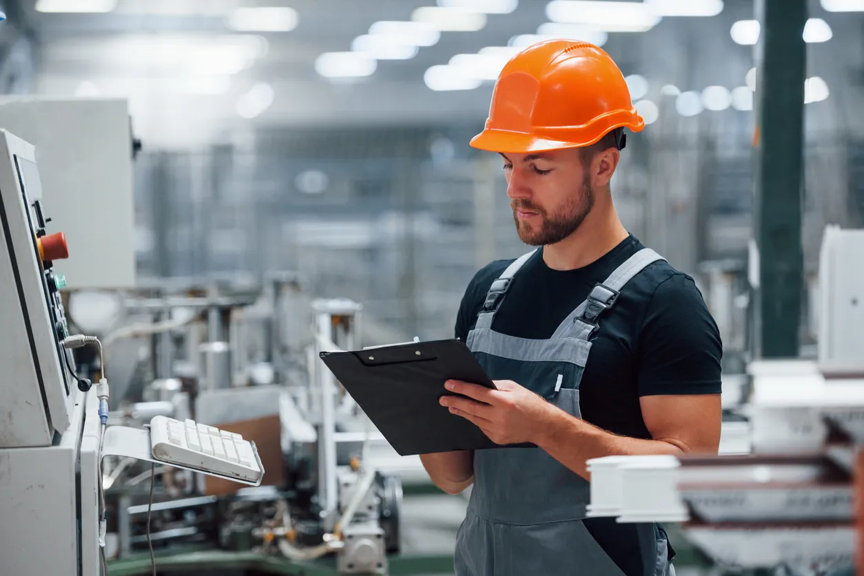 Mira el equipo y toma notas. Trabajador industrial en interior en fábrica. Joven técnico con sombrero duro naranja