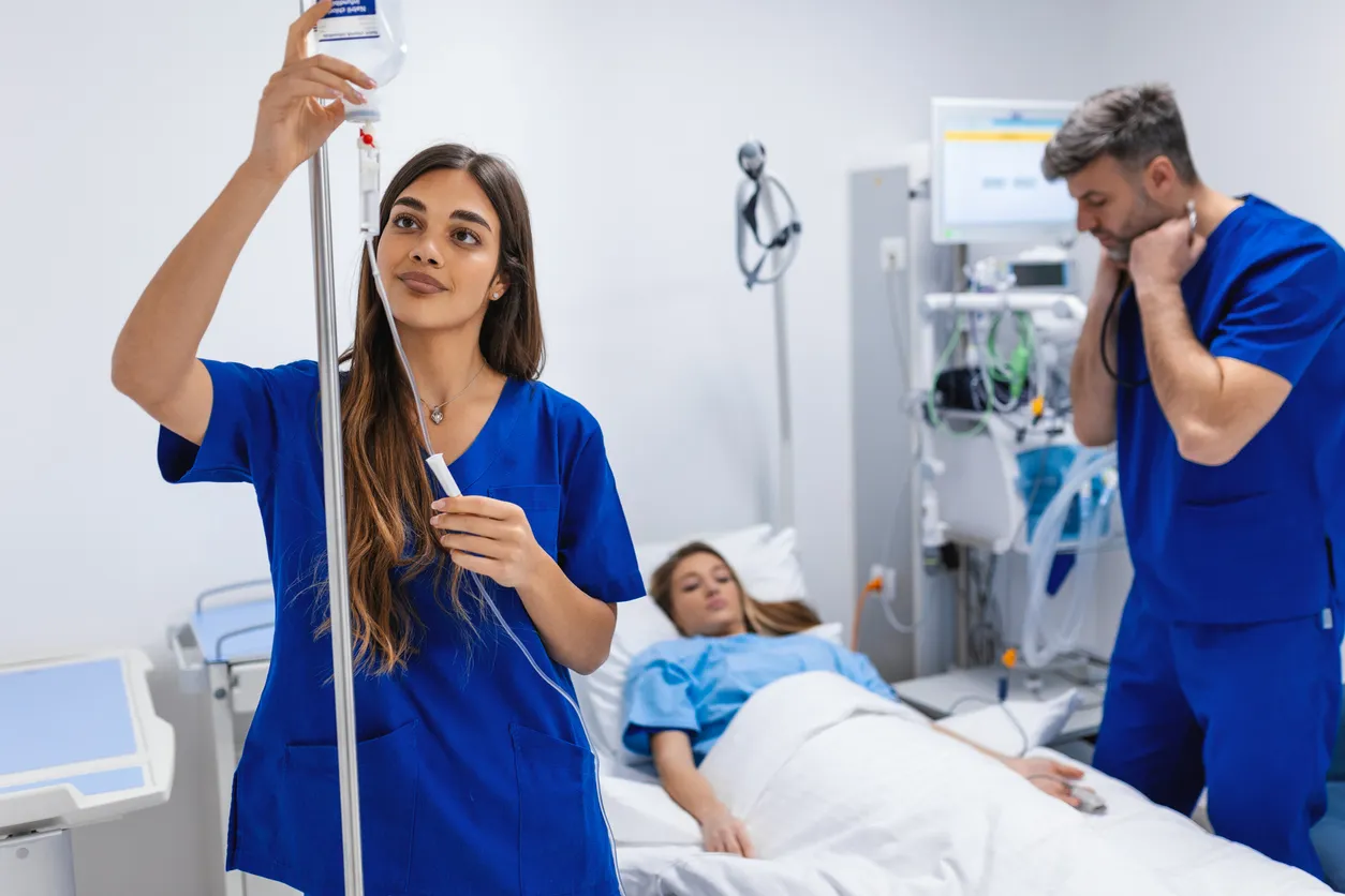 Una joven enferrmera vestida con scrub médico azul, coloca el gotero intravenoso en la habitación del hospital.