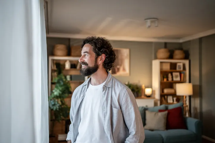 Hombre barbudo sonriente mirando por la ventana en casa