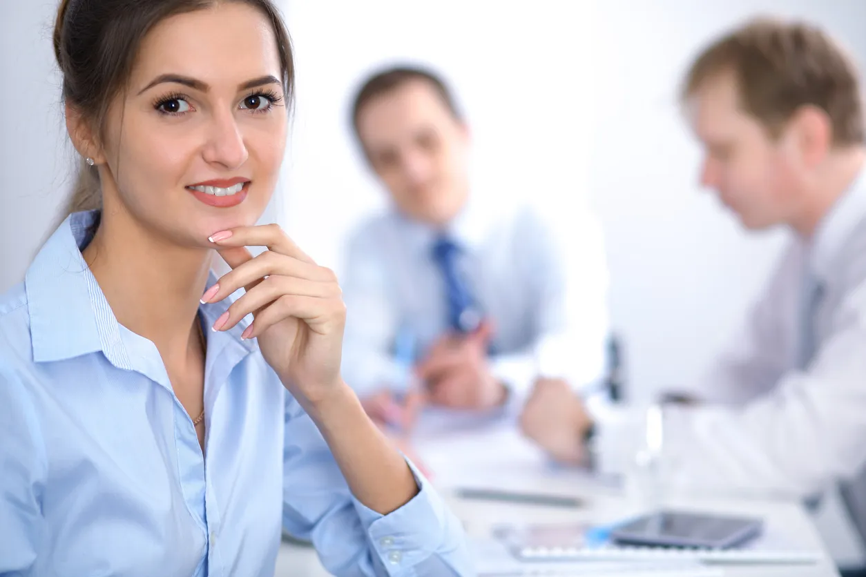 Mujer con uniforme en color azul en oficina con dos compañeros.al fondo. 