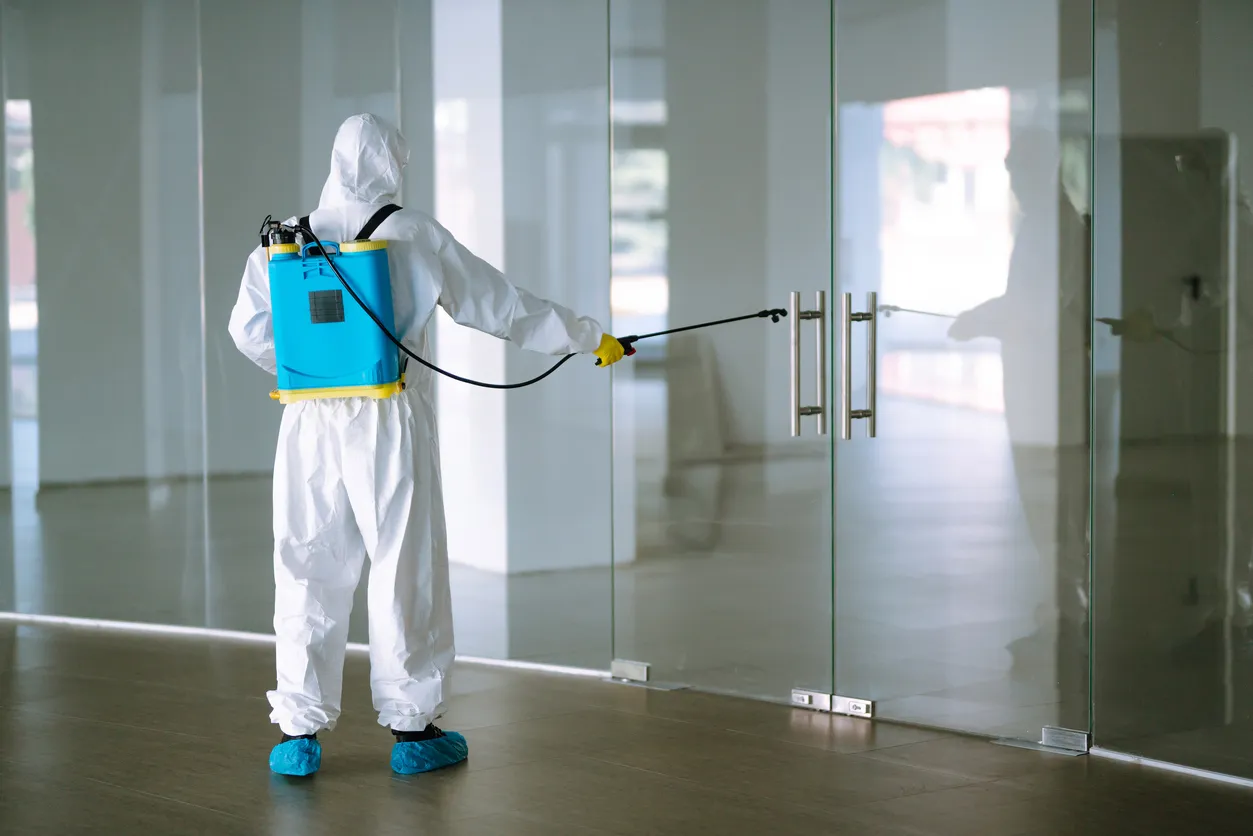 Hombre con traje hazmat, guantes de protección y gafas utilizan equipo pulverizador desinfectar en la oficina. 