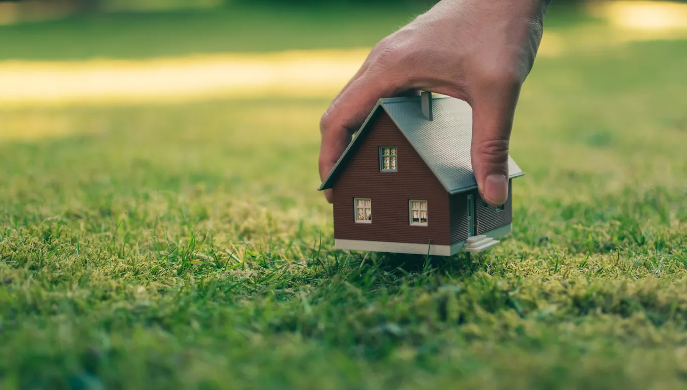 Concepto de venta de una casa. Una mano sostiene una casa modelo por encima del prado verde