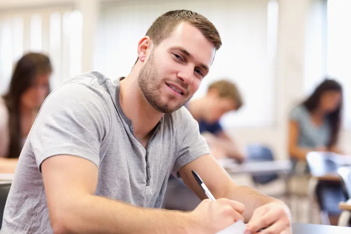Hombre joven sonriendo y sentado en un escritorio estudiando 