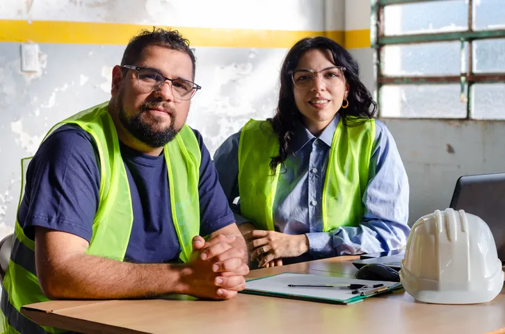 Dos jovenes sonrientes y con chalecos de seguridad sentados en una área de estudio