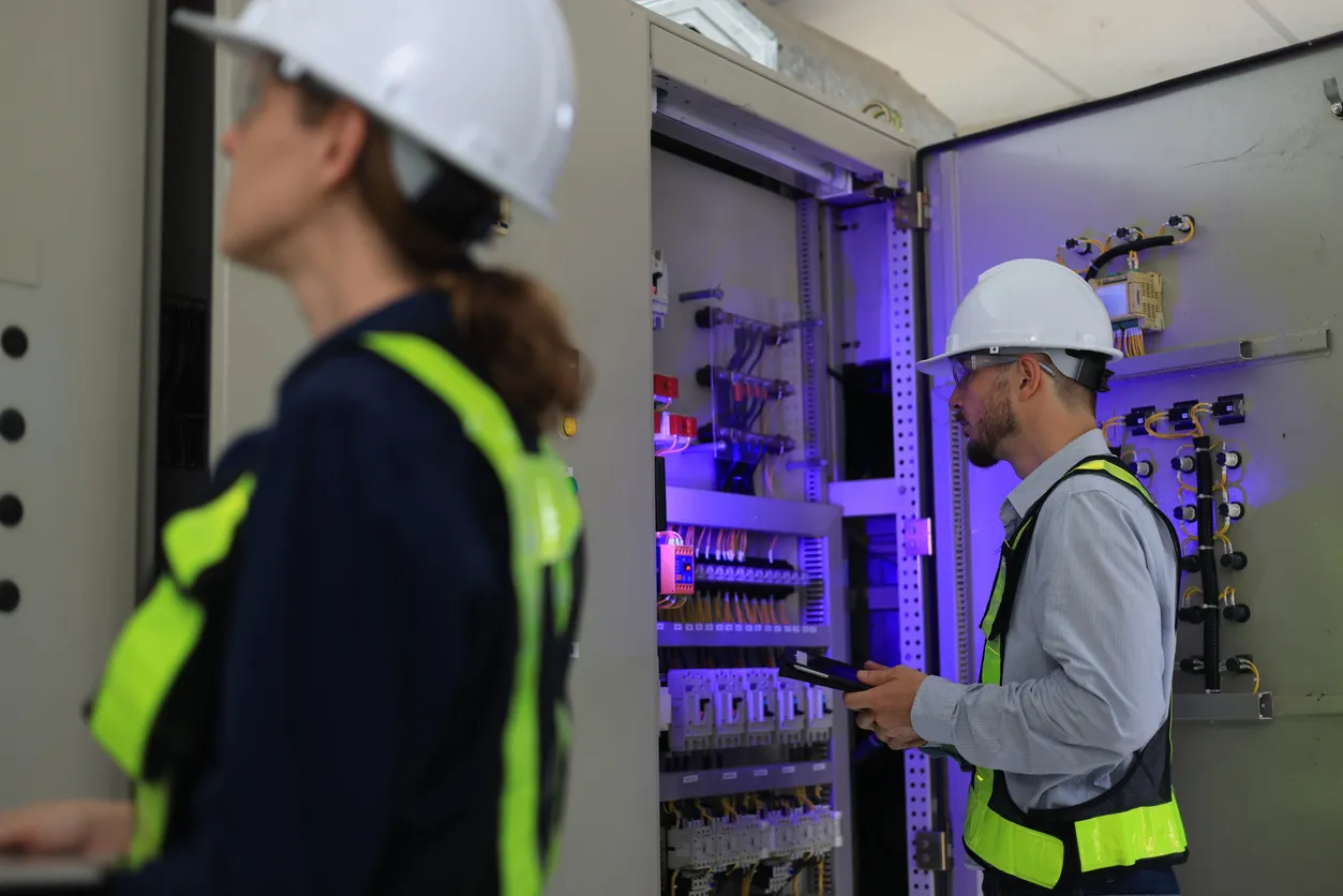 Los ingenieros electricistas de servicios industriales están uno al lado del otro, intercambiando ideas frente al tablero de 